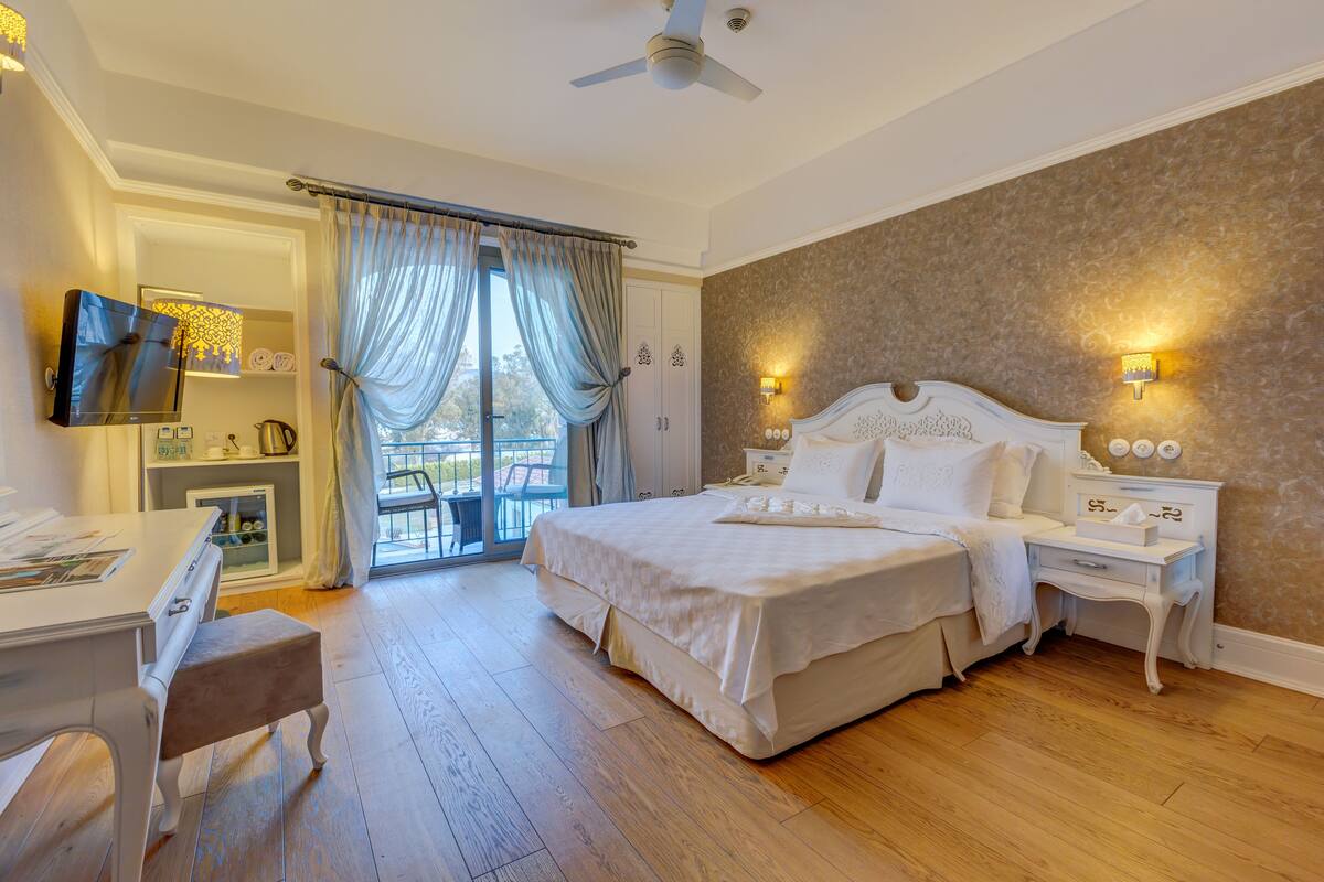 A bedroom with a large bed, a desk, a chair, a TV, and a balcony with curtains.