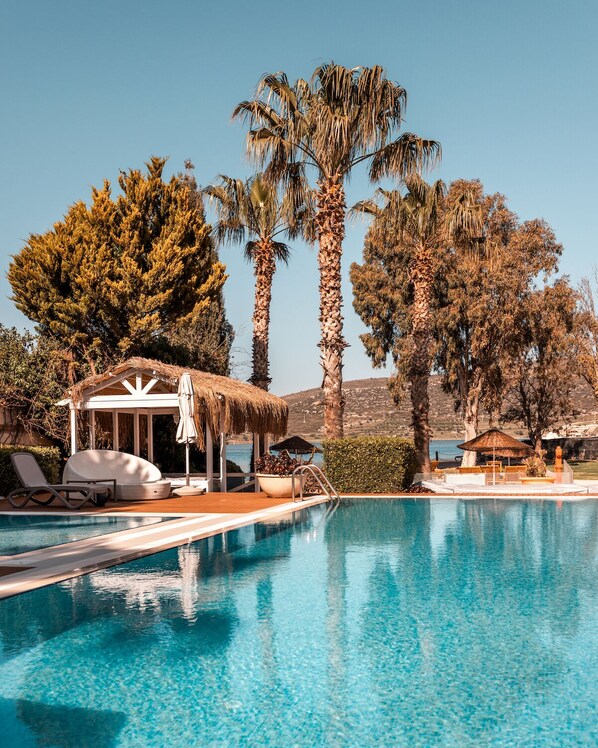 A pool area with lounge chairs, a thatched-roof structure, and tall palm trees. - Antmare Hotel (Cesme)