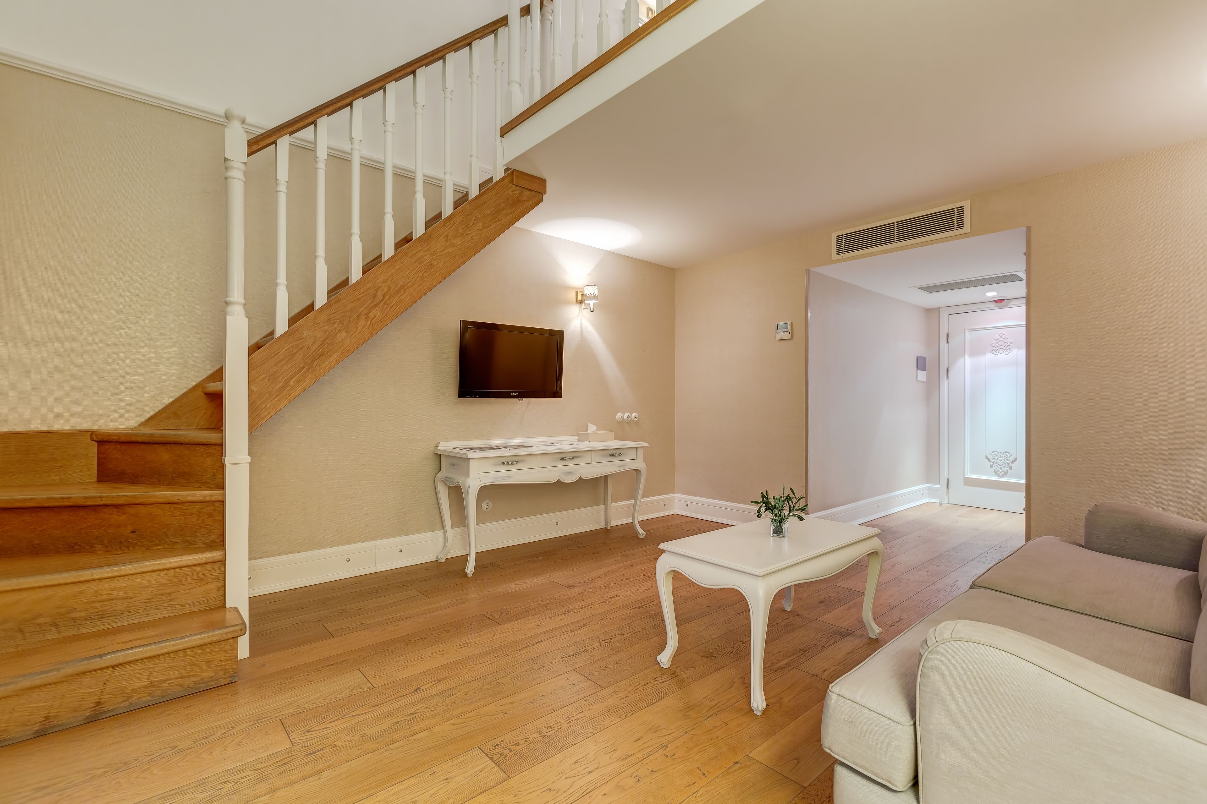 Una sala de estar con una escalera de madera, una consola blanca, una mesa de centro blanca y un sofá color beige.