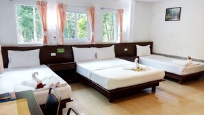 Ein Hotelzimmer mit drei Einzelbetten, einem hölzernen Kopfteil, einem kleinen Schreibtisch mit Stuhl und Blick auf Grün durch die Fenster.