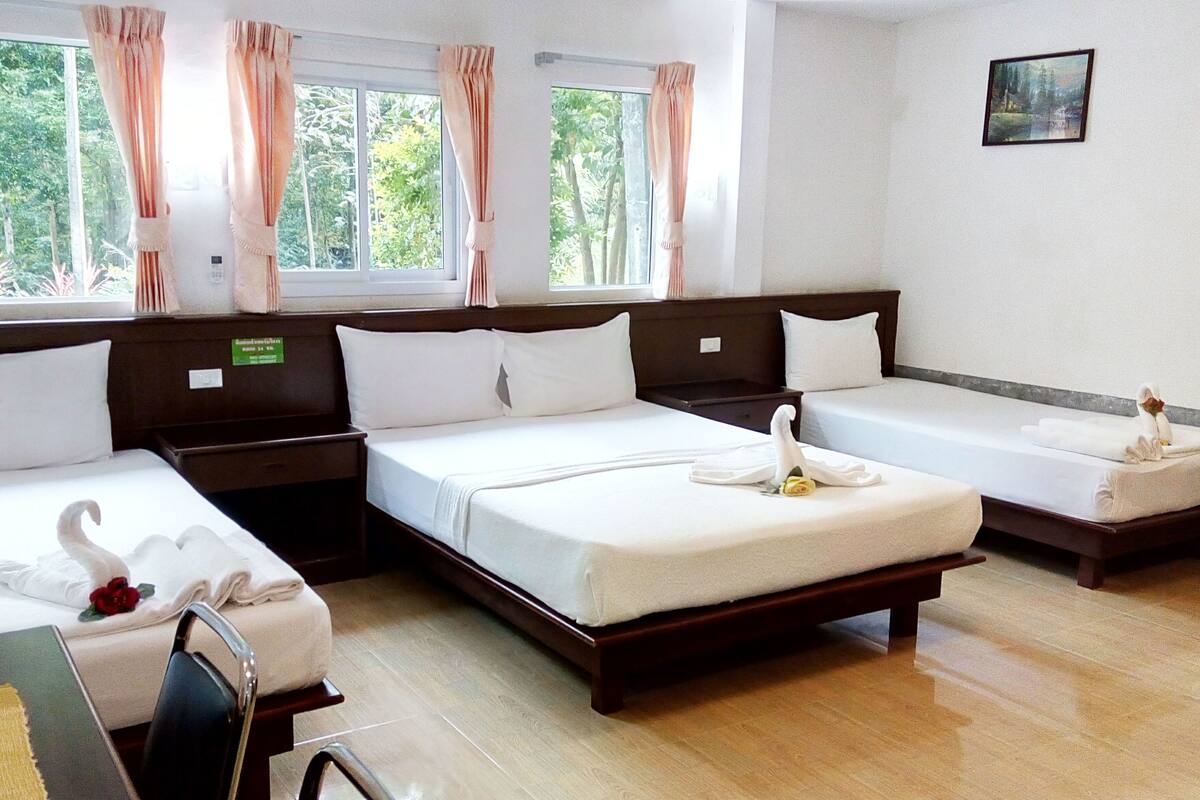 Habitación de hotel con tres camas individuales, cabecera de madera, un escritorio pequeño con una silla y vistas a zonas verdes por las ventanas.