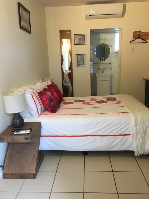 A hotel room with a bed, a nightstand, a lamp, a mirror, and a bathroom visible through a glass door.