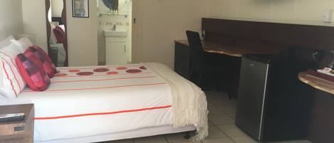 A hotel room with a bed, a desk, a chair, a TV, and a minibar.
