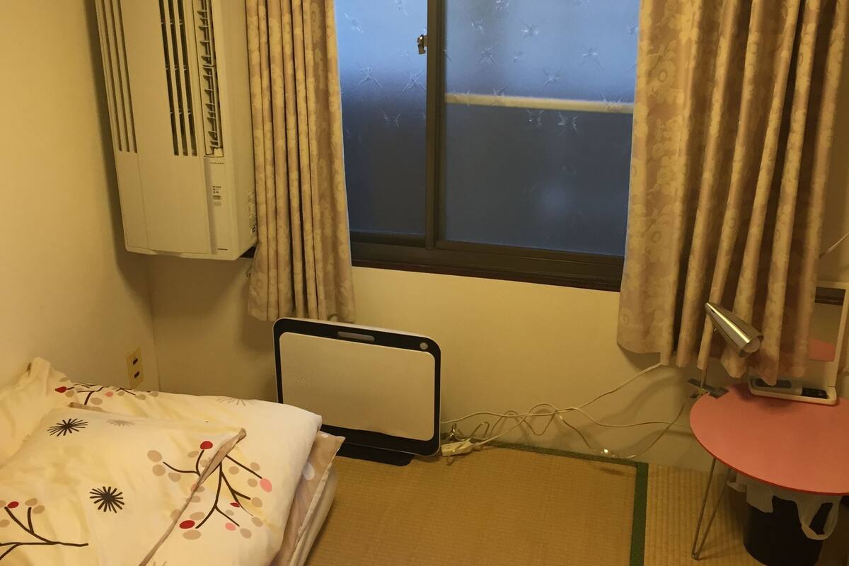 Habitación de hotel con una cama, una mesita de noche, una pequeña mesa con lámpara, una ventana con cortinas y un equipo de aire acondicionado.