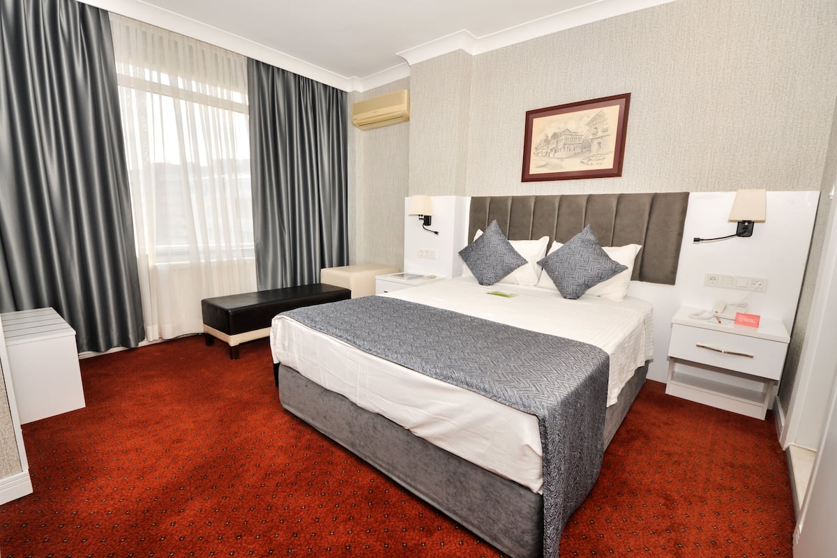 A hotel room with a large bed, bedside tables, a bench, a wall-mounted air conditioner, and a framed picture on the wall.
