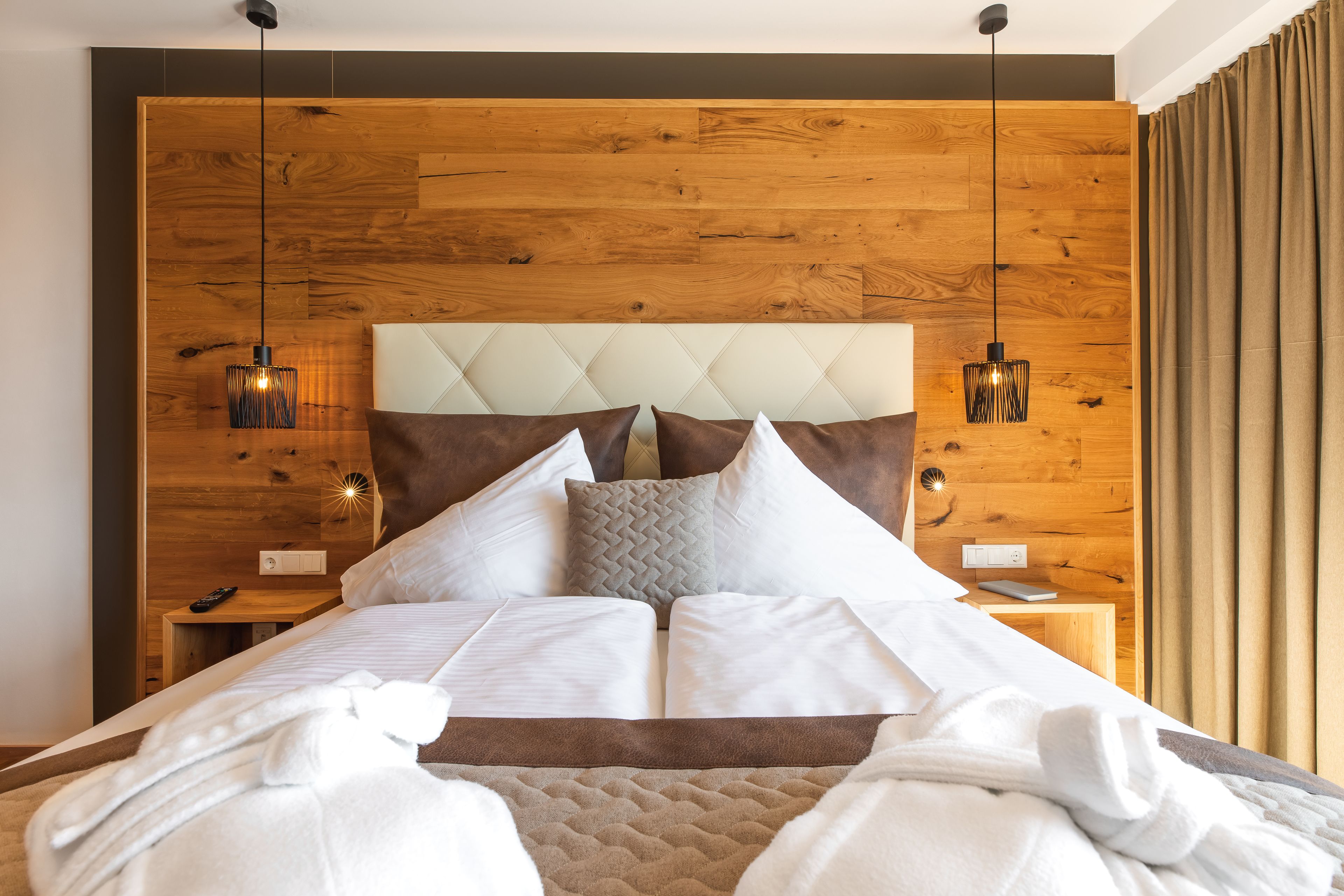 A modern hotel room with a wooden headboard, a large bed with white linens, two bedside tables, and a nightstand.