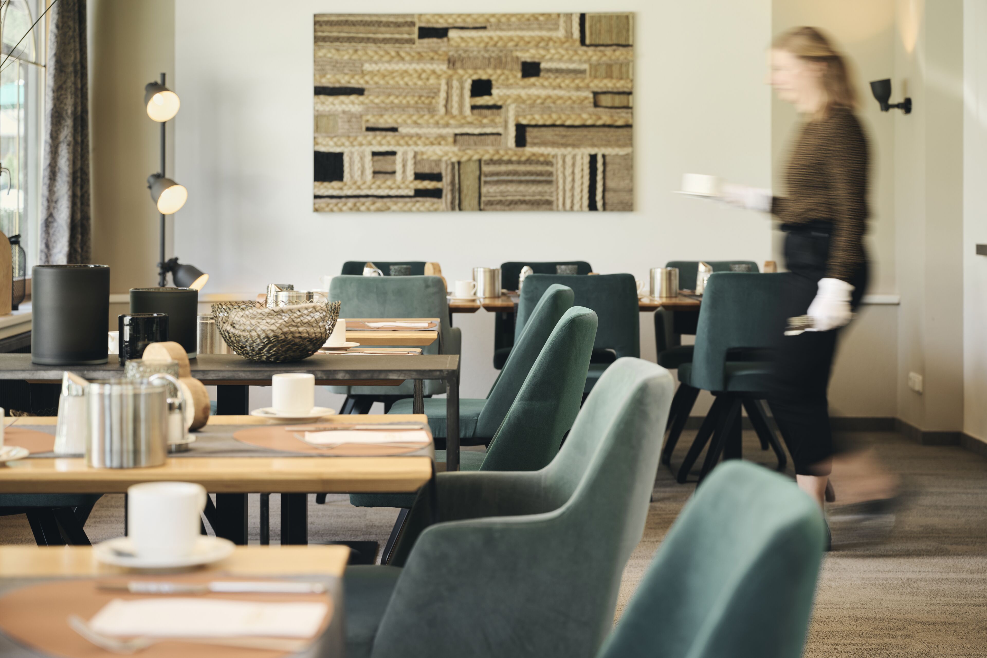 Un área de comedor moderna con mesas de madera y sillas tapizadas de verde, una cesta tejida y una obra de arte abstracta colgada en la pared.