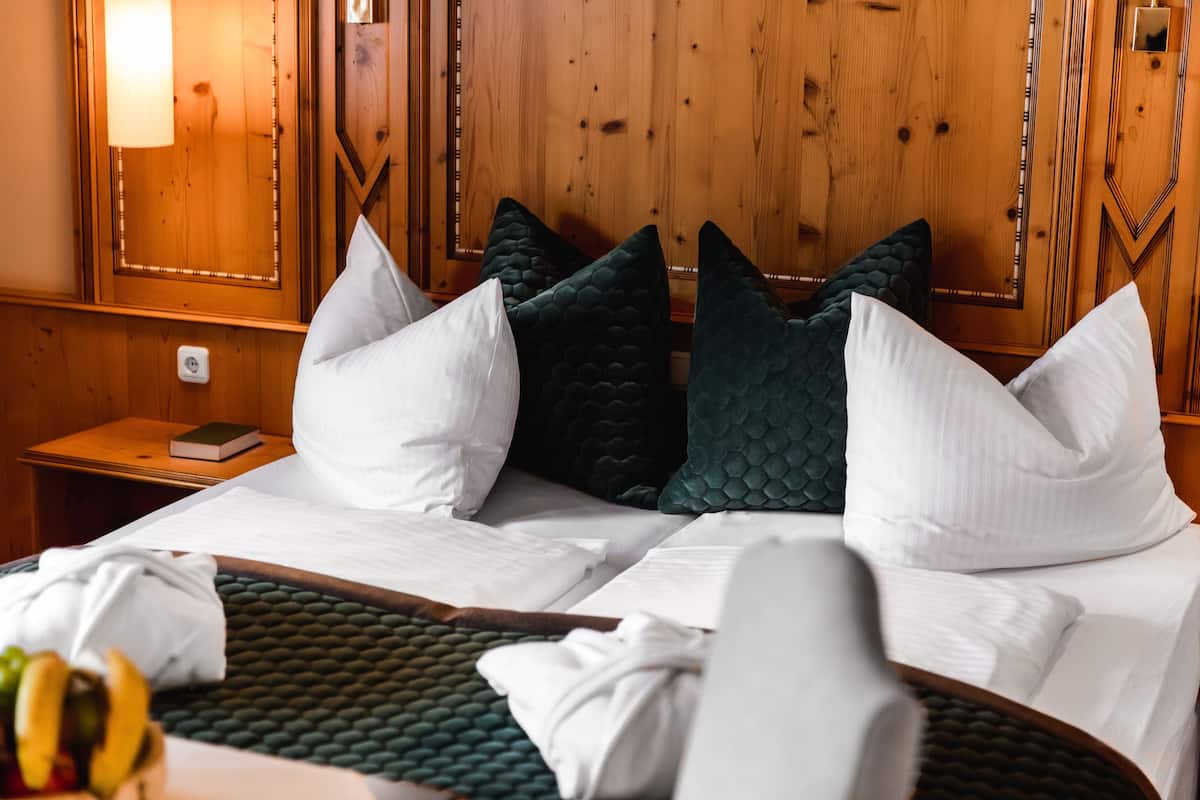 Habitación de hotel con cabecera de madera, dos camas con almohadas blancas y verde oscuro, una mesita de noche con un libro y una lámpara, y un frutero a la izquierda.