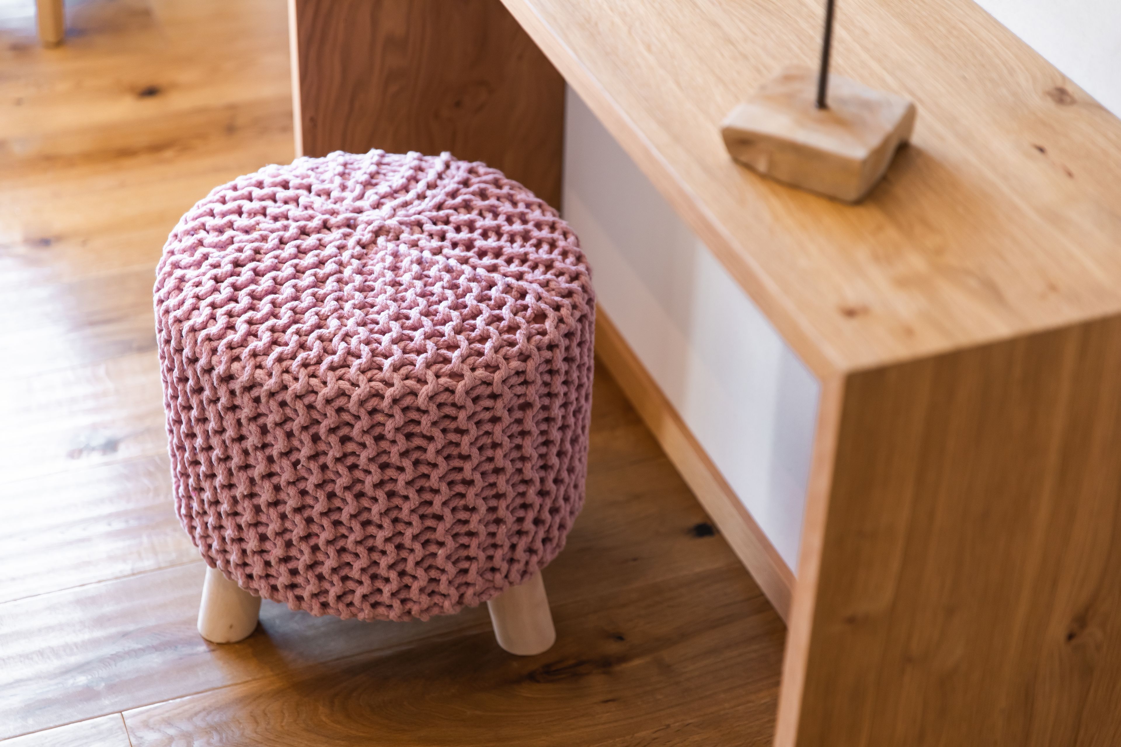 Un otomán rosa de punto con patas de madera sobre piso de madera.
