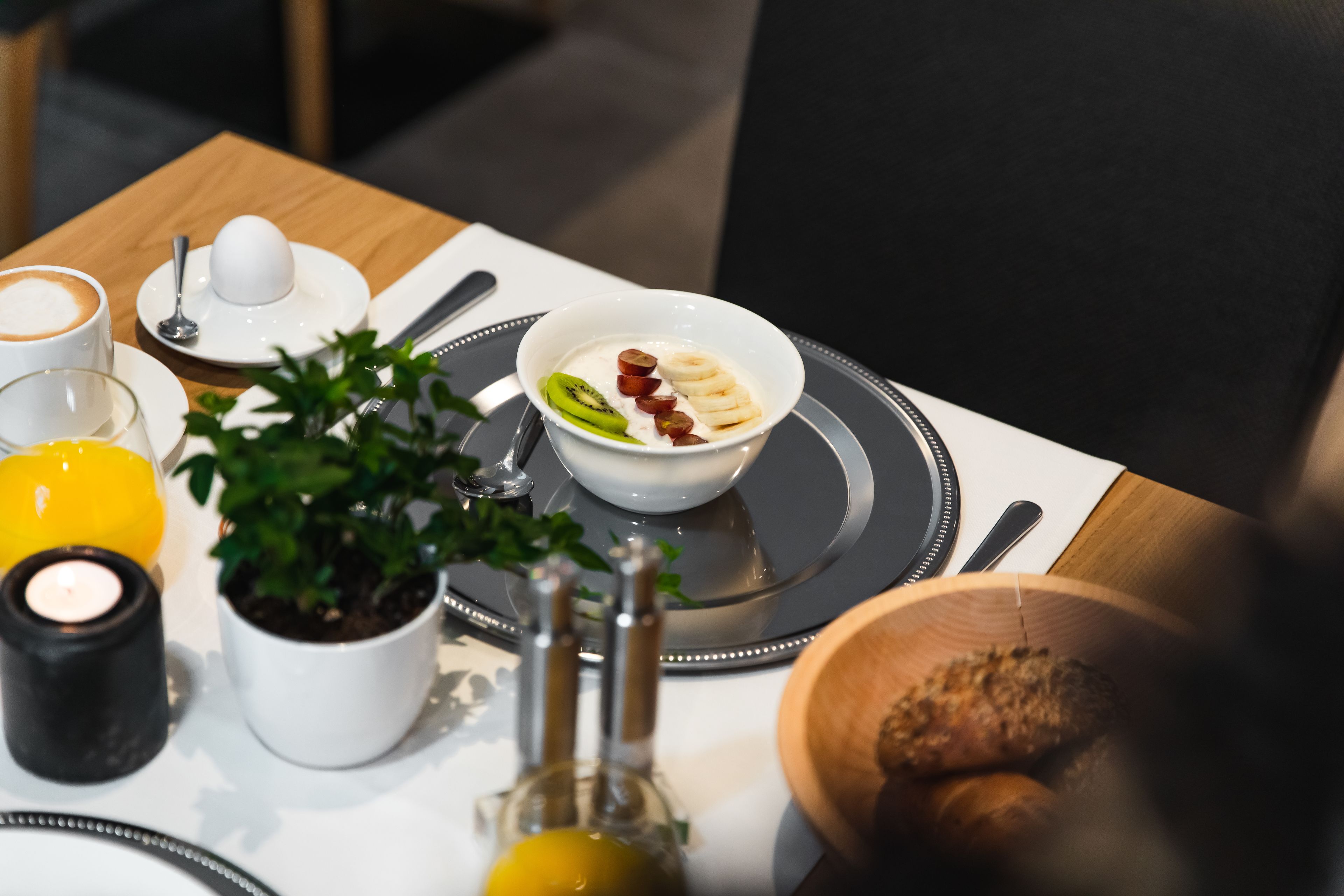 Un desayuno con un tazón de yogur cubierto de kiwi, uvas y rodajas de plátano, una taza de café, un vaso de jugo de naranja y una vela sobre una mesa.