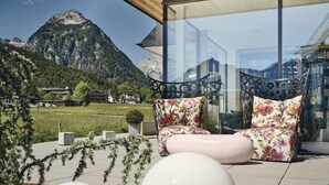 A modern outdoor seating area with a mountain view, featuring a glass wall and a metal chair with floral cushions. - Hotel Auszeit (Eben am Achensee)