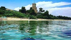 A coastal scene with clear turquoise water, a sandy beach, and a tower-like structure surrounded by greenery.