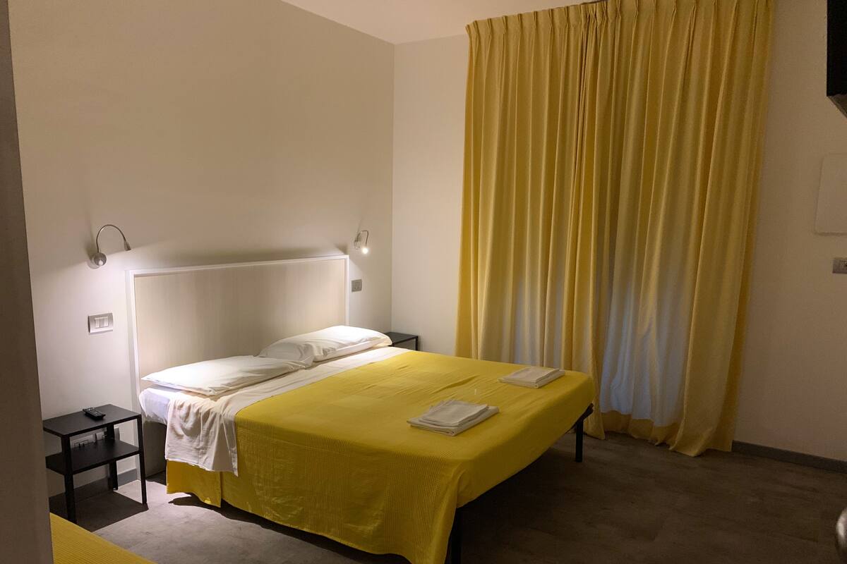 Un letto rifatto con una coperta gialla, un comodino con una lampada e una finestra con tende gialle.