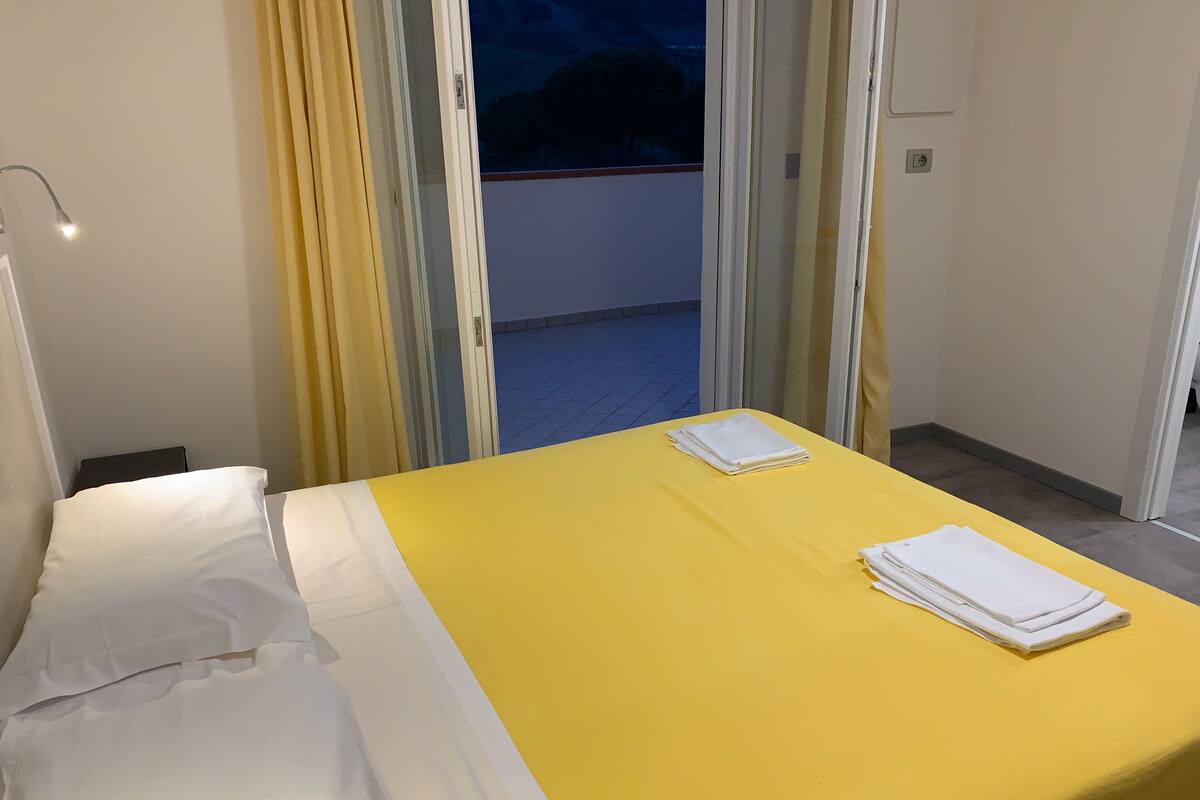 Una camera d'albergo con un letto, una coperta gialla e una finestra con tende.