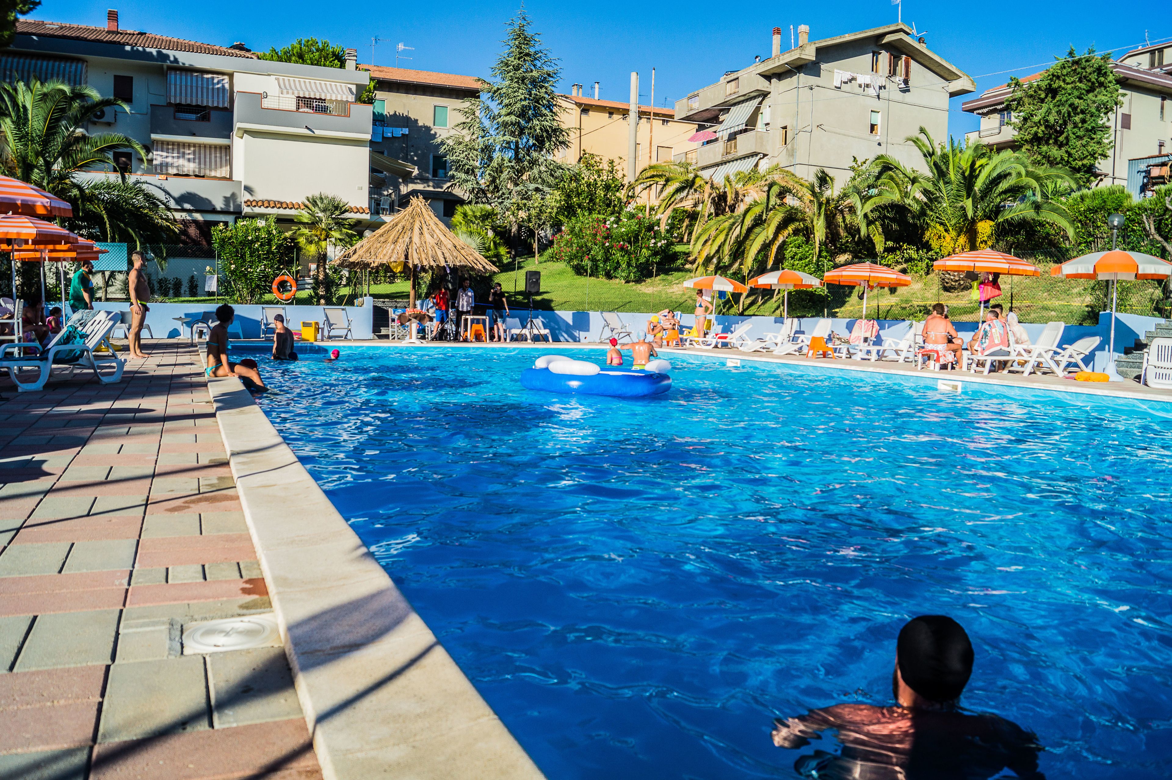 Piscina con sillas de descanso y sombrillas, rodeada de edificios y palmeras.