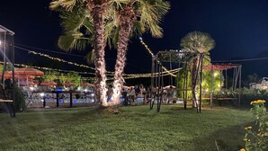 An outdoor dining area with string lights, palm trees, and tables under umbrellas.
