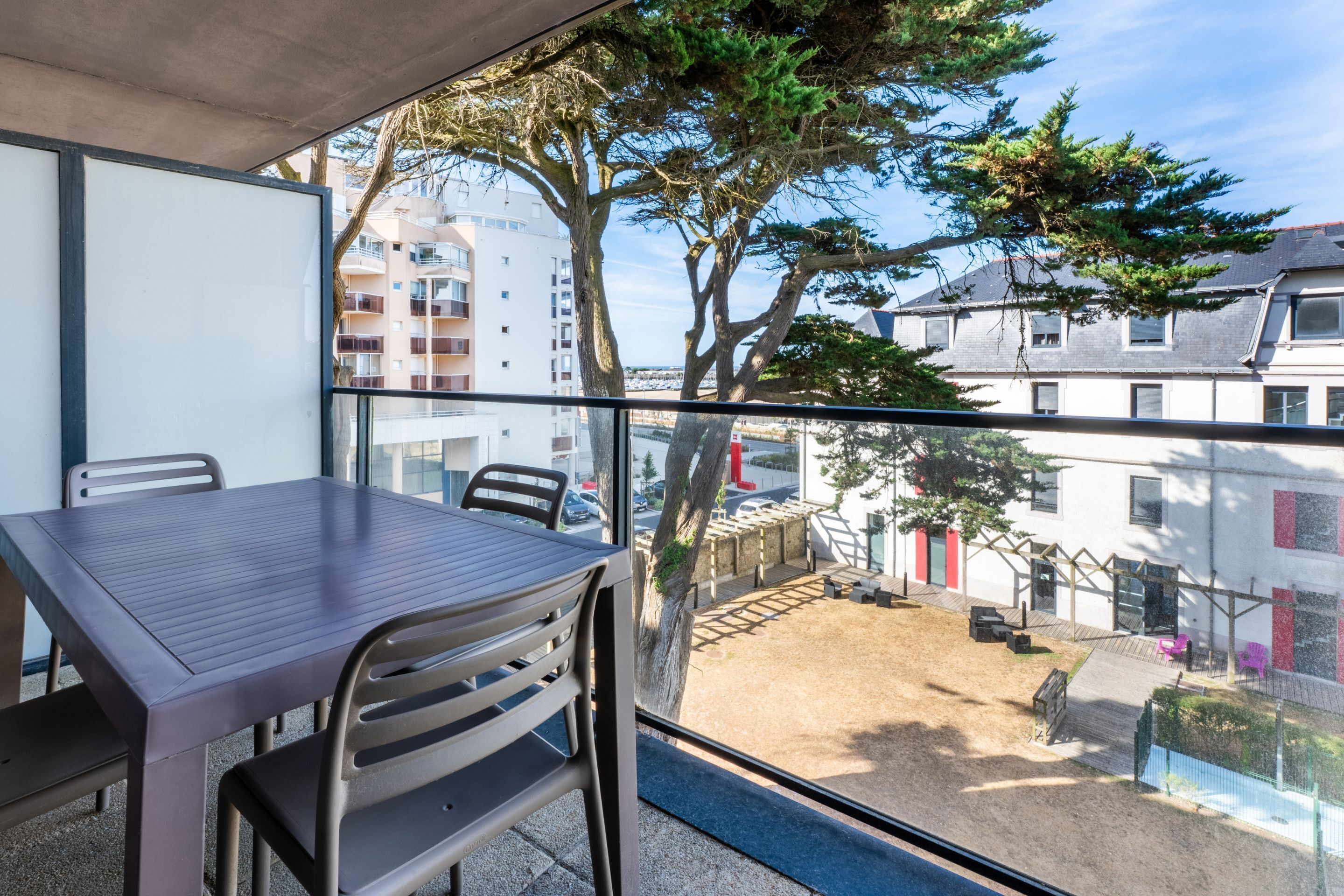 A balcony with a table and chairs, overlooking a courtyard with trees and buildings.