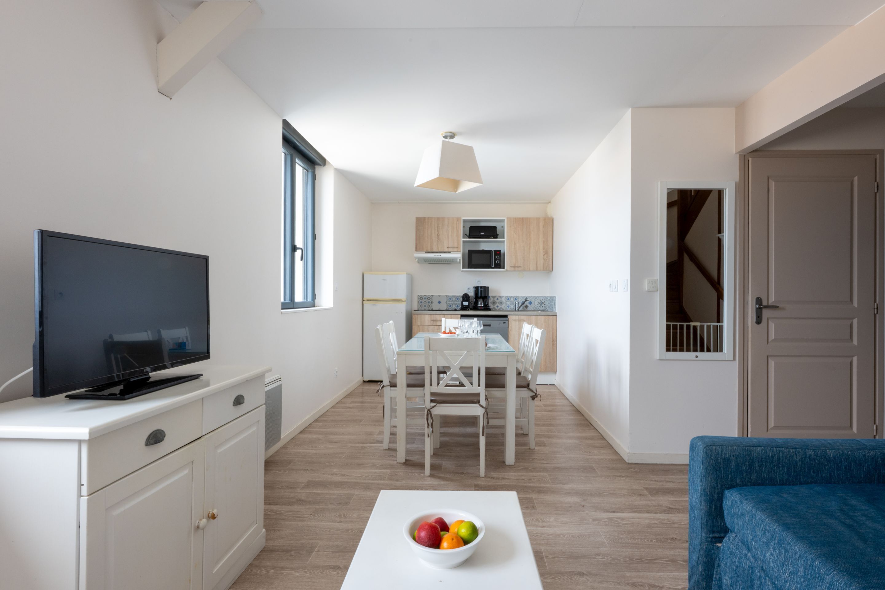 Un espace de vie compact comprenant un téléviseur, une table à manger et un saladier de fruits posé sur une table blanche.