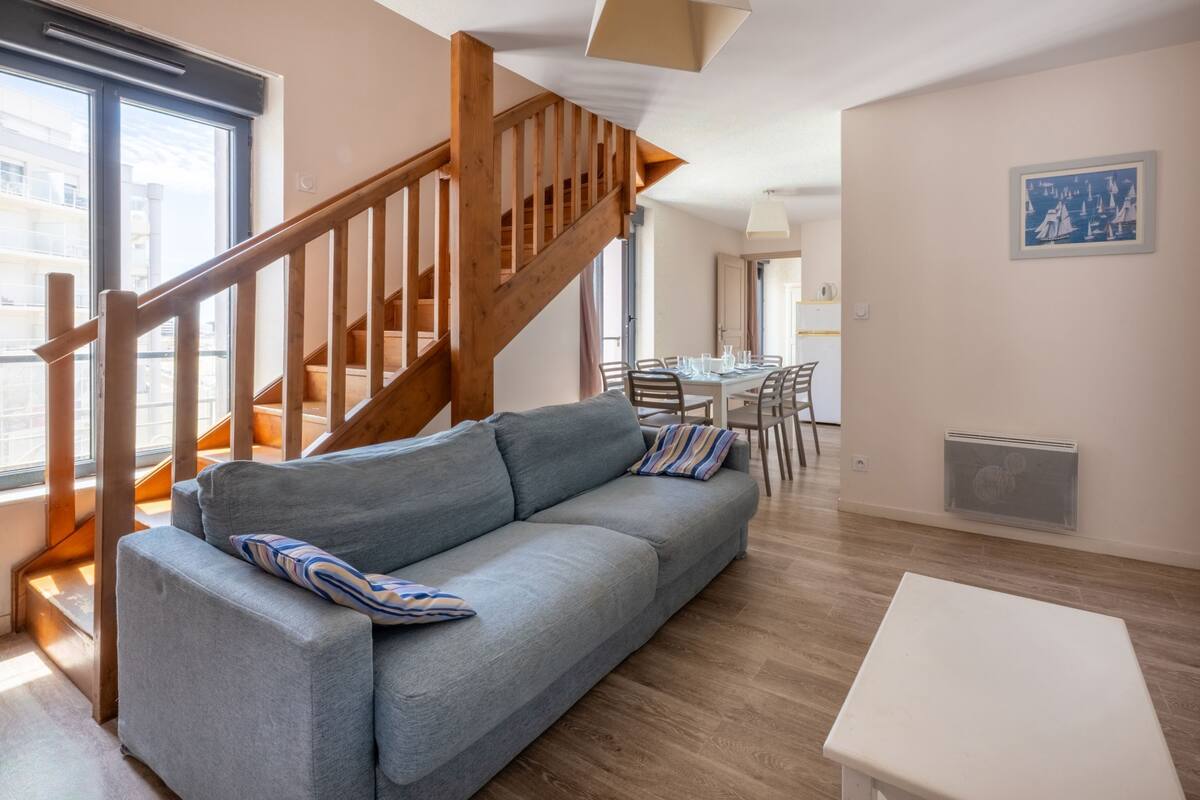 A modern living room with a grey sofa, wooden staircase, and a dining area in the background.
