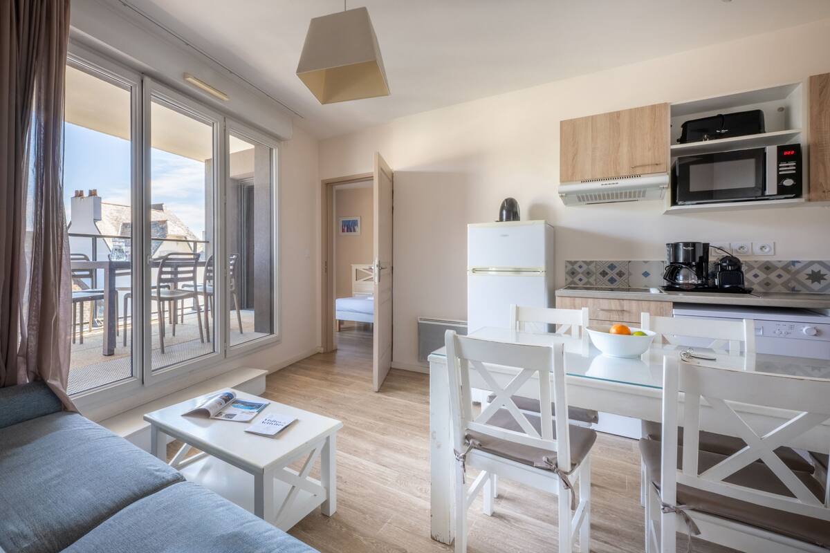 A modern kitchen with white cabinets, a microwave, and a coffee maker. There is a dining table with chairs and a window overlooking a balcony.