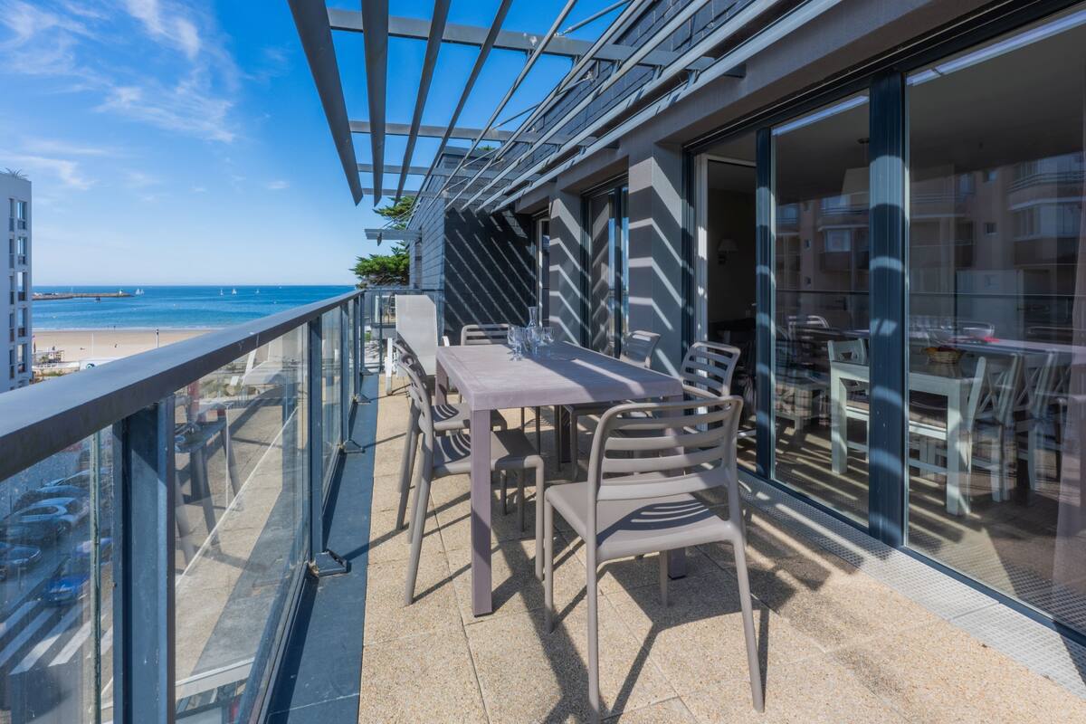 An outdoor balcony with a table and chairs, overlooking a beach and ocean.