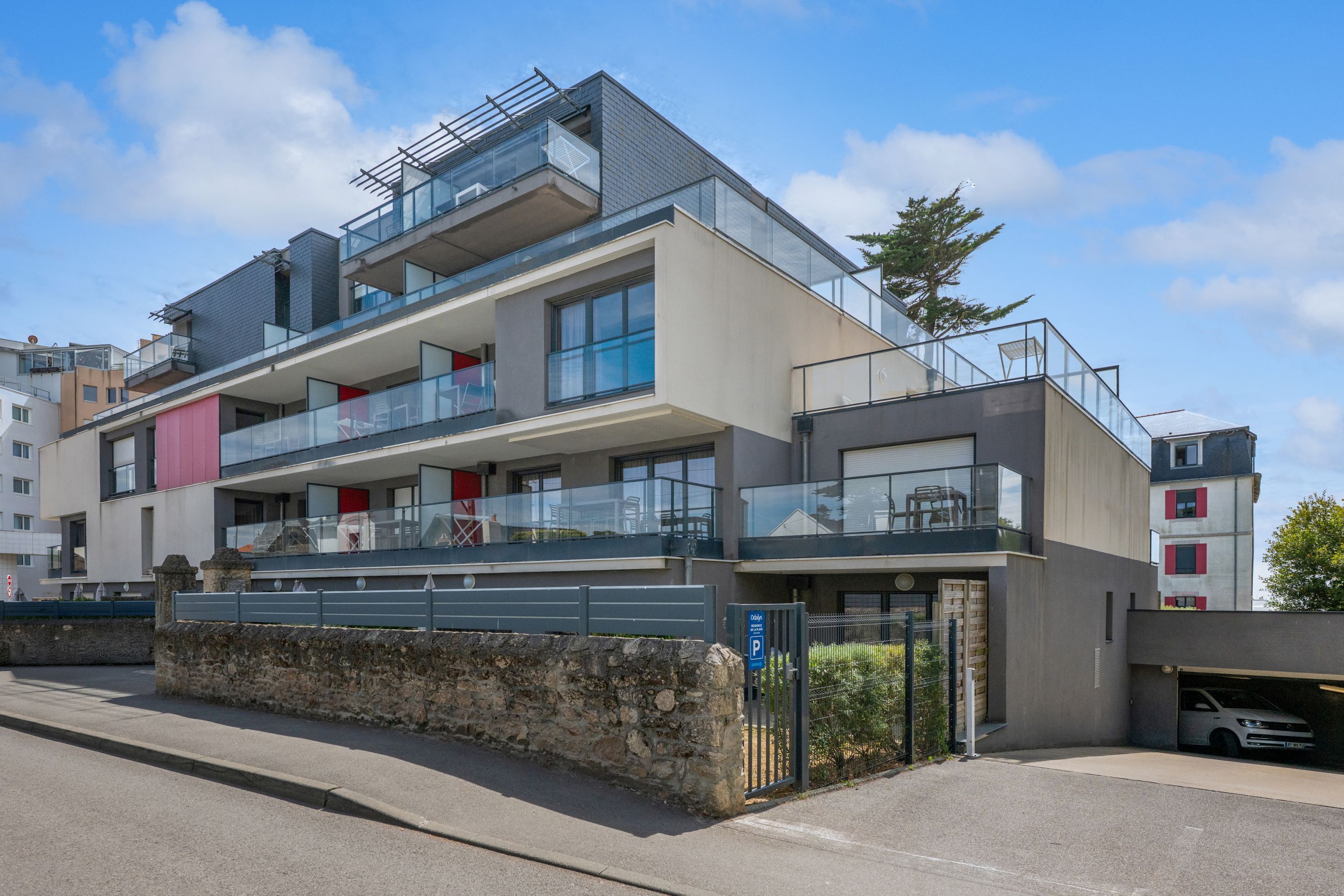 A modern multi-story residential building with balconies, large windows, and a parking area.