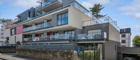 Un immeuble résidentiel moderne à plusieurs étages, doté de balcons, de grandes fenêtres et d’un espace de stationnement.
