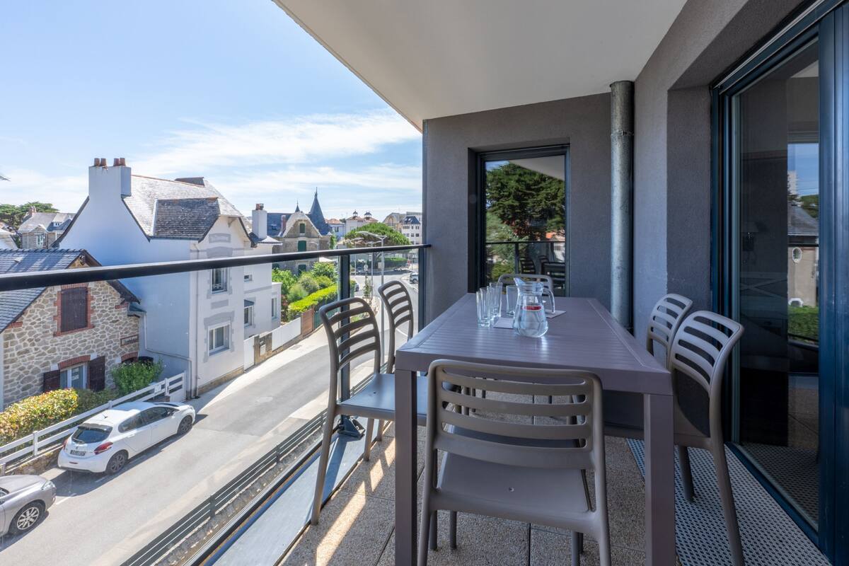 A balcony with a table and chairs, overlooking a street with parked cars and buildings.