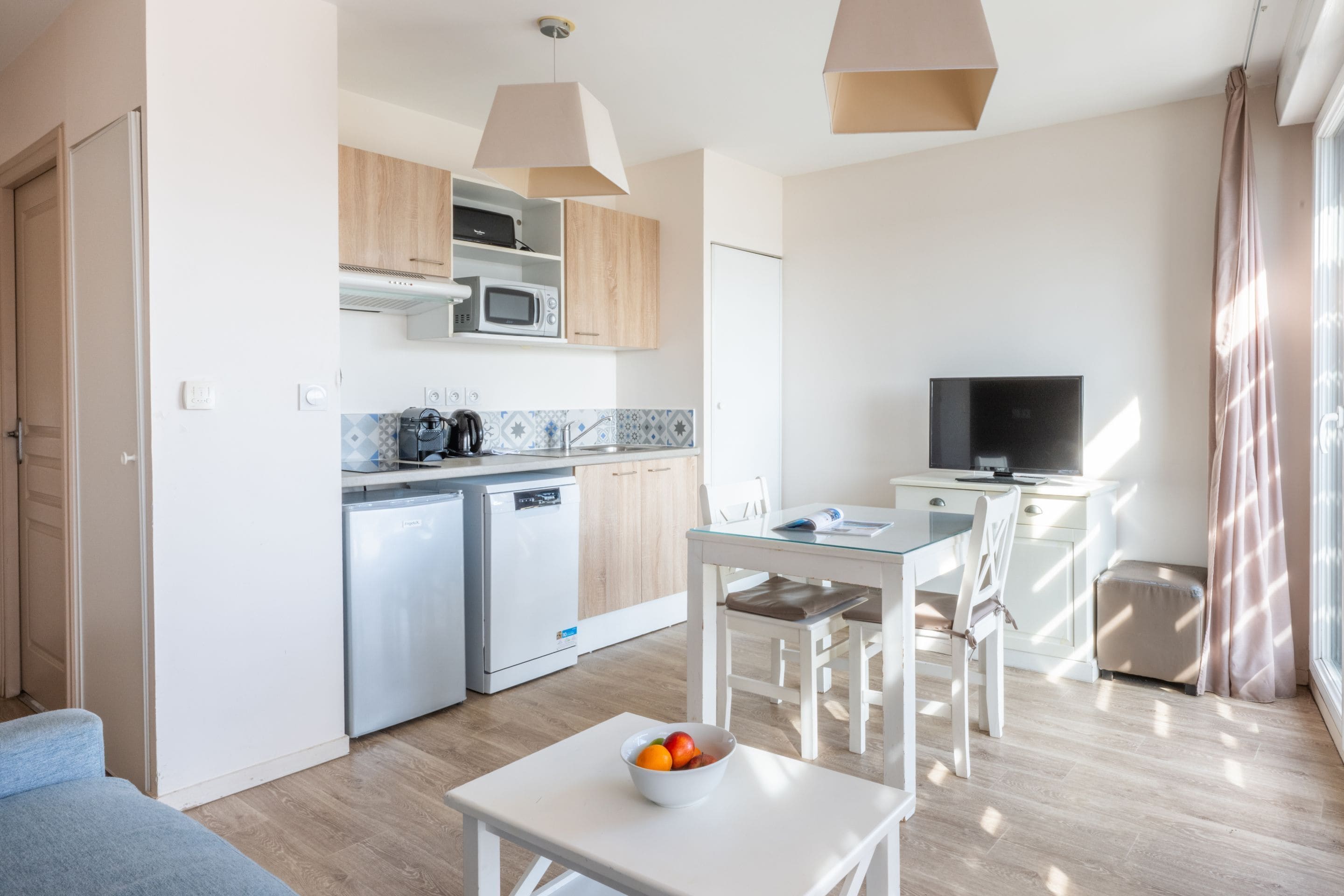 A modern kitchen with white cabinets, a microwave, and a dishwasher. There is a dining area with a table and chairs, and a TV on a stand.