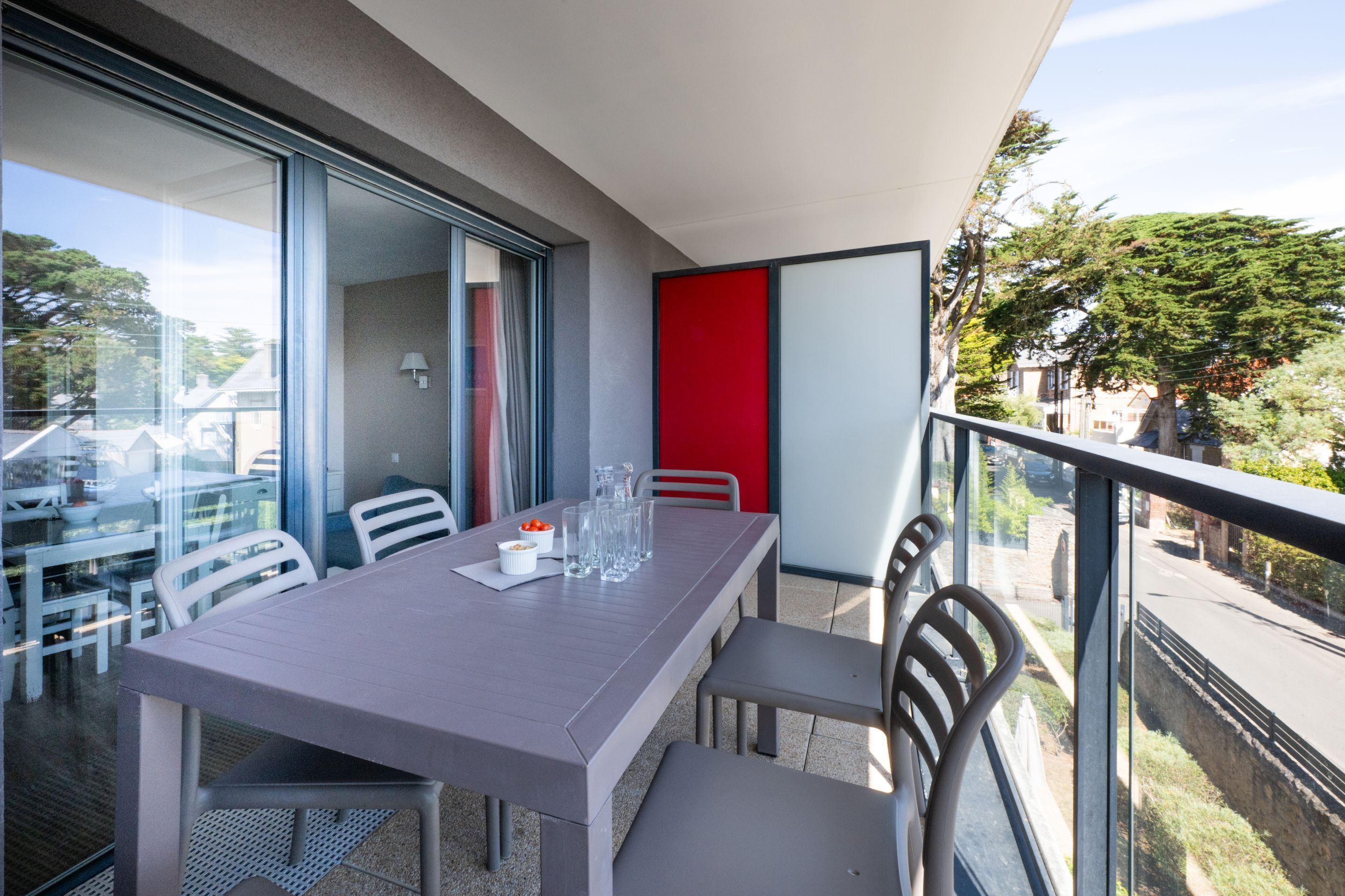 Ein Balkon mit Tisch und Stühlen, eine rote Akzentwand und Blick auf die Straße sowie Bäume.