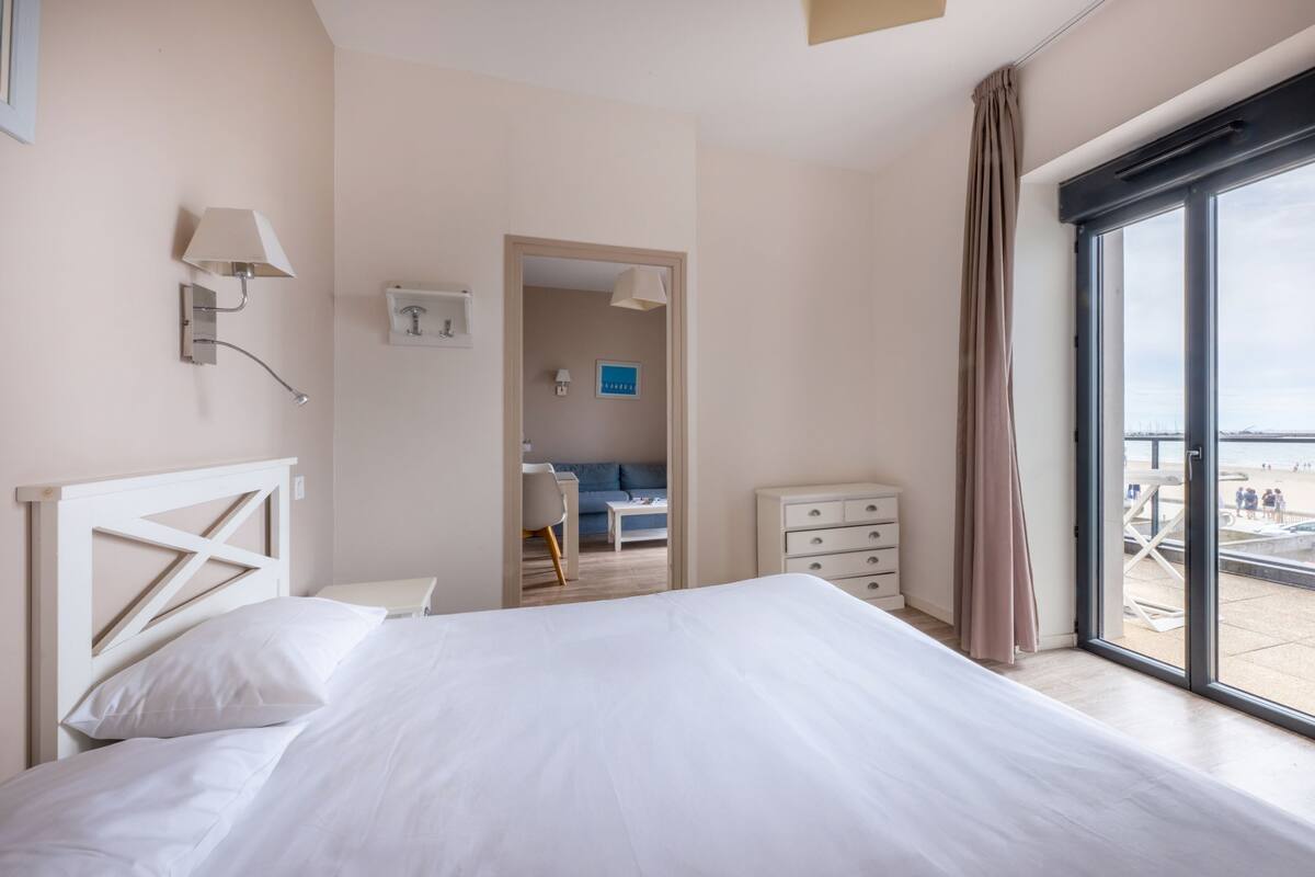 A hotel room with a bed, a desk, a dresser, and a view of the beach.