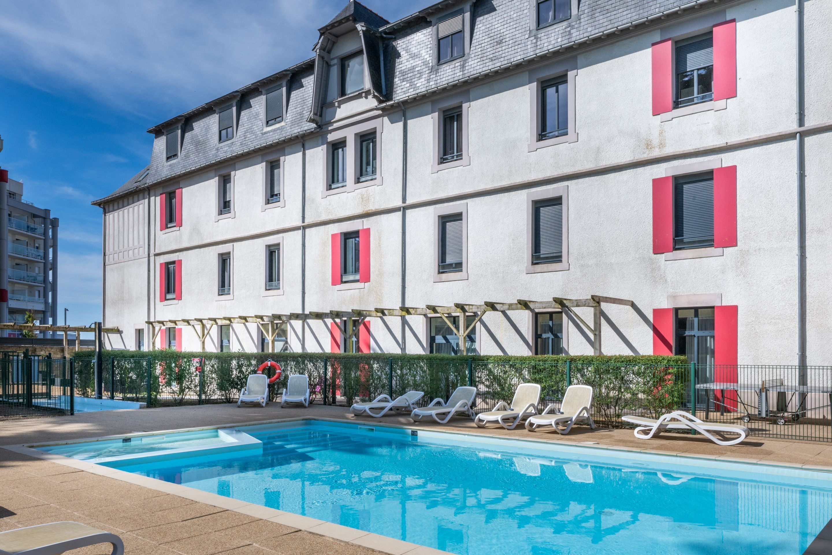 Una piscina con sillas de descanso, un edificio con contraventanas rojas y un cielo azul despejado.