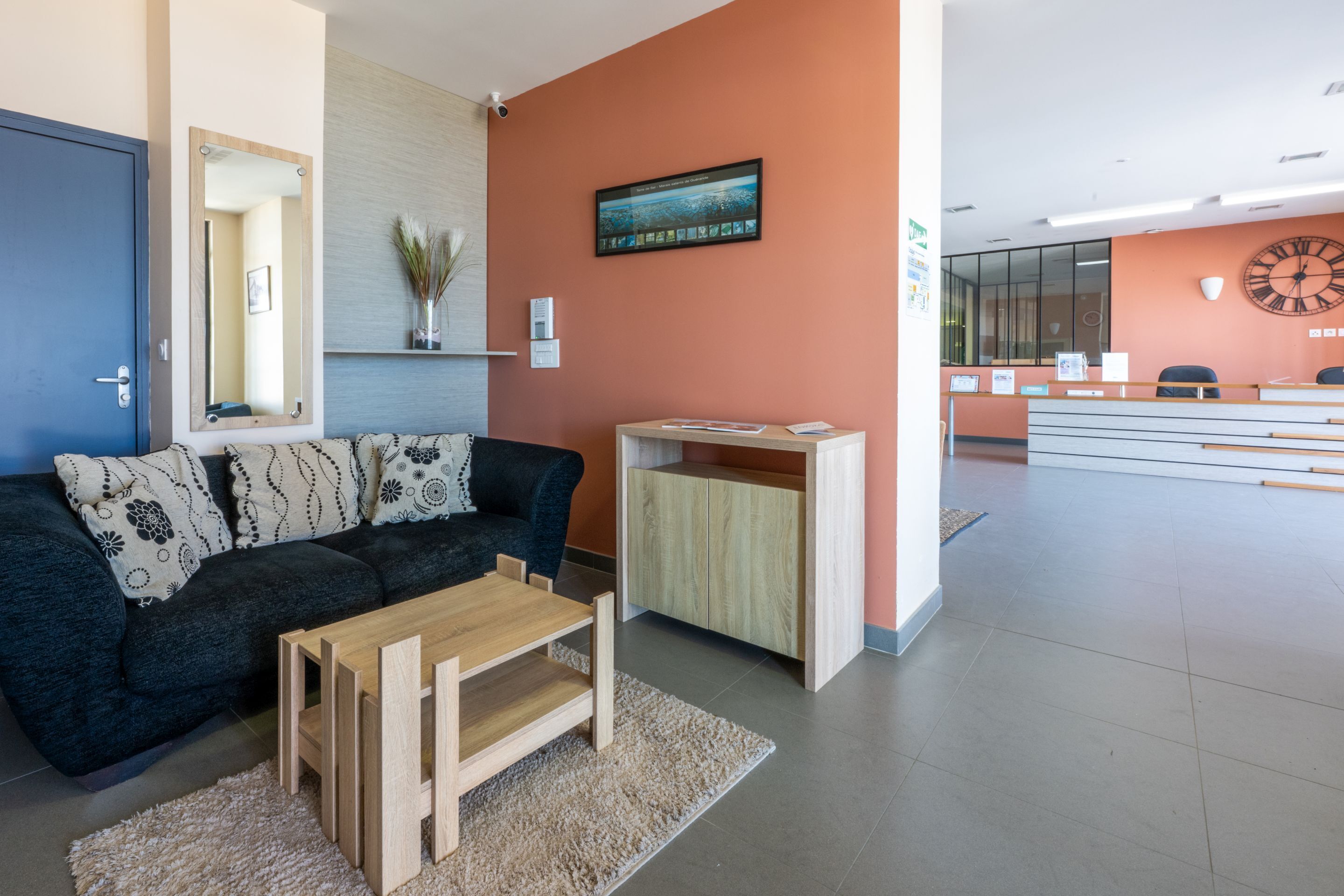 Ein moderner Empfangsbereich mit einem Holztisch, einem schwarzen Sofa mit gemusterten Kissen, einem Spiegel und einer Wanduhr.
