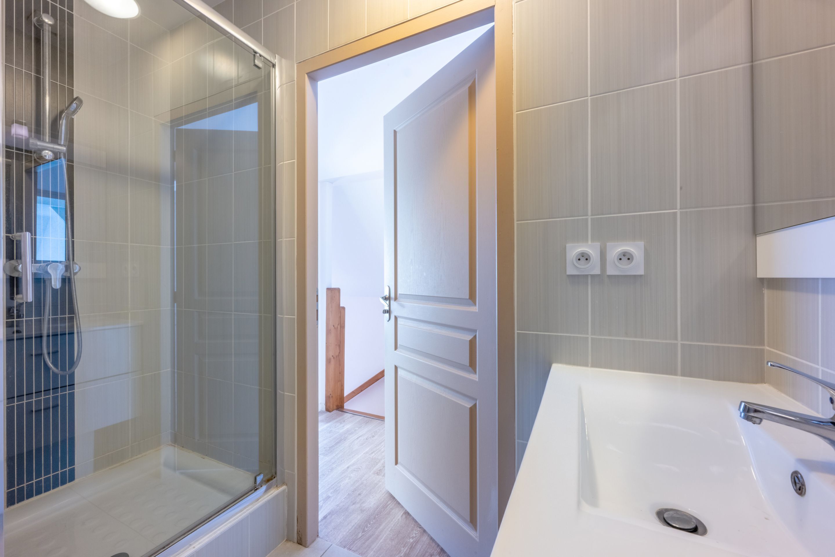 Een moderne badkamer met een douchecabine van glas, een witte wastafel en een houten deur die naar een andere kamer leidt.