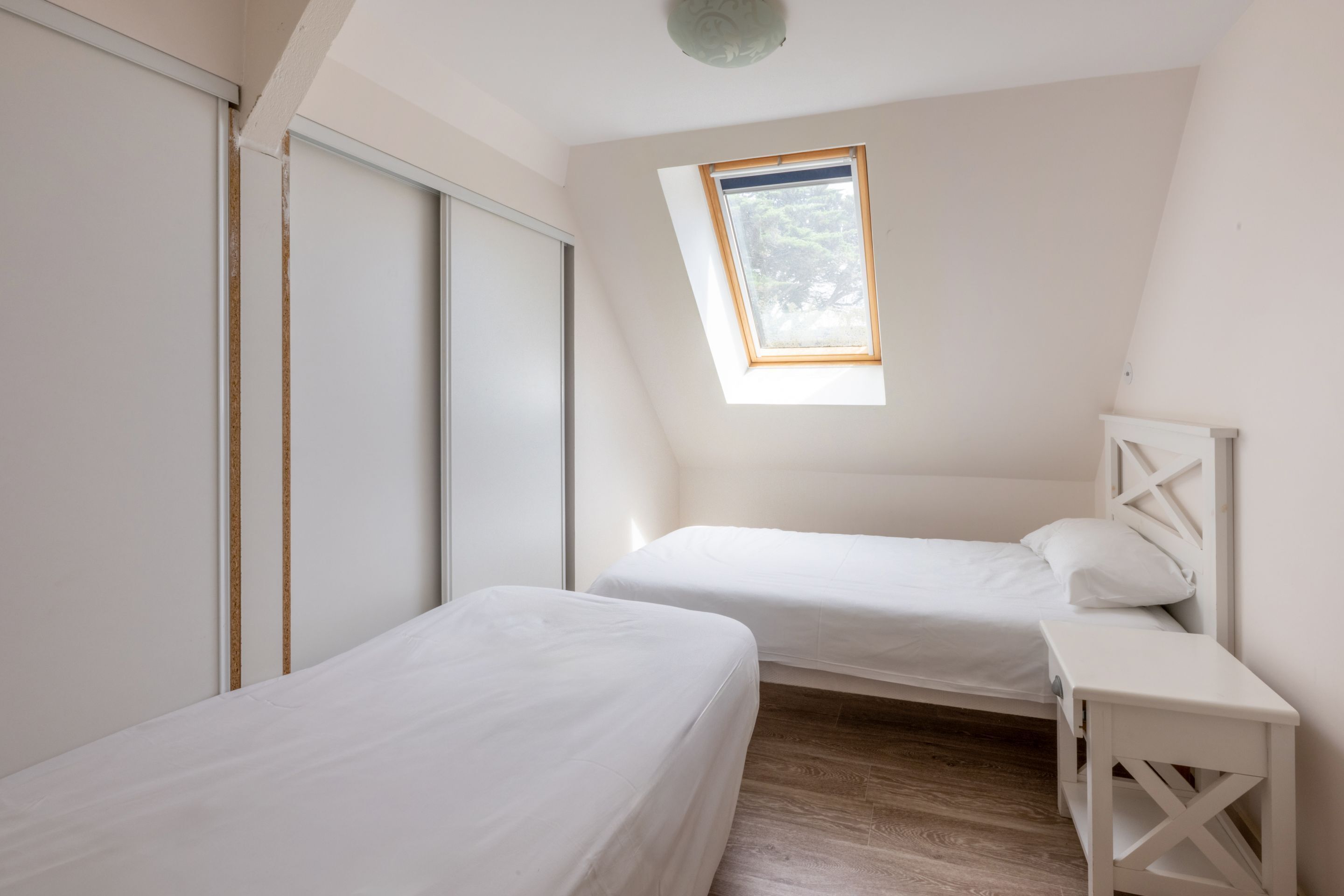 Una habitación pequeña y blanca con dos camas, un tragaluz y suelo de madera.