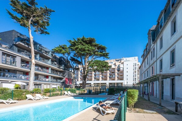 Ein Poolbereich mit Liegestühlen, einer Pergola und mehrstöckigen Gebäuden im Hintergrund.