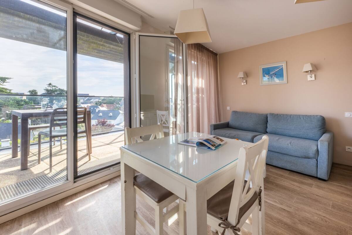 A modern living room with a glass table, a blue sofa, and a sliding glass door leading to a balcony.