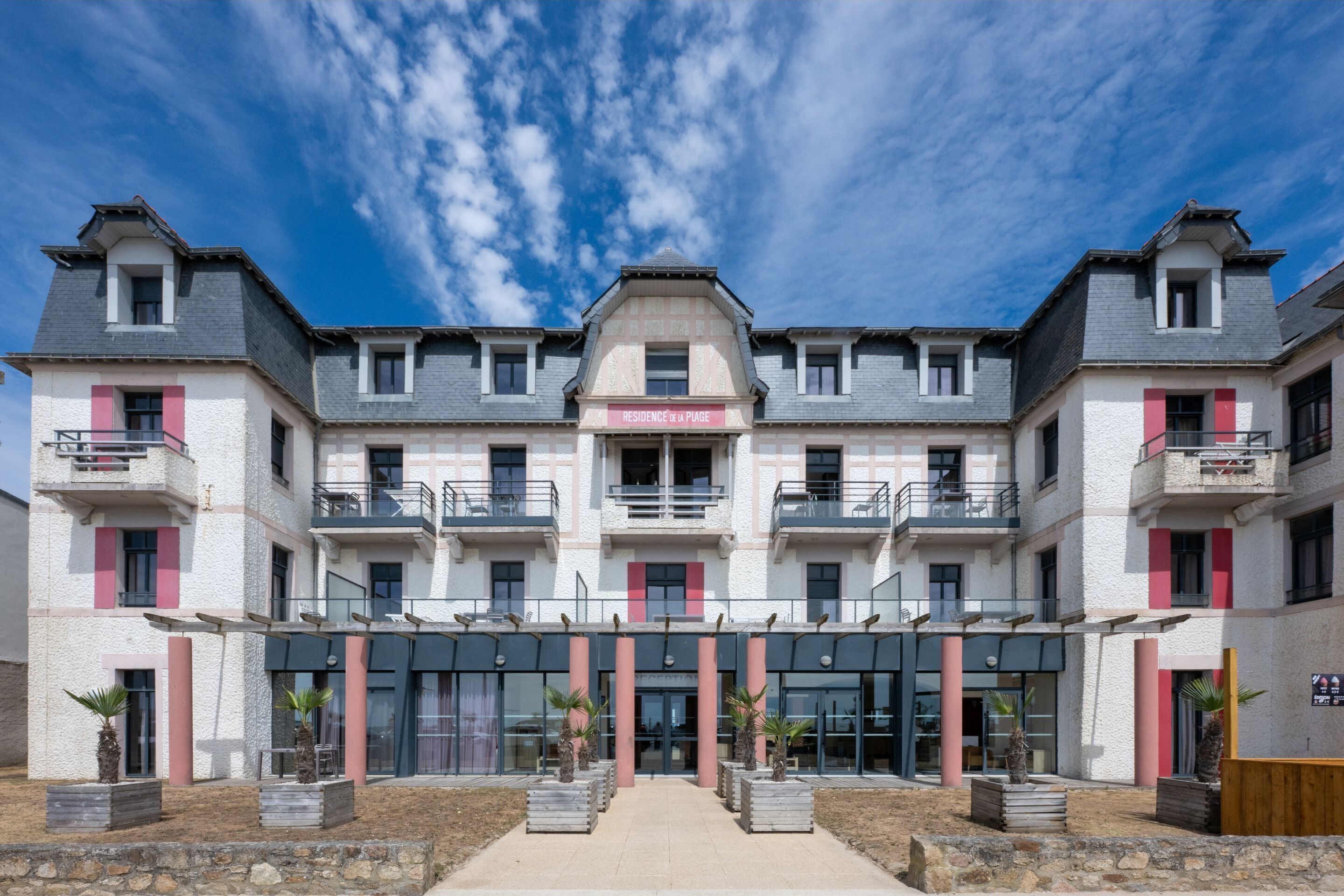 A multi-story hotel with a central entrance, balconies, and potted plants.