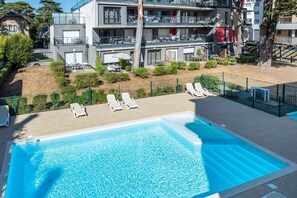 Ein Schwimmbecken mit Liegestühlen, eine umzäunte Terrasse und ein mehrstöckiges Gebäude mit Balkonen im Hintergrund.