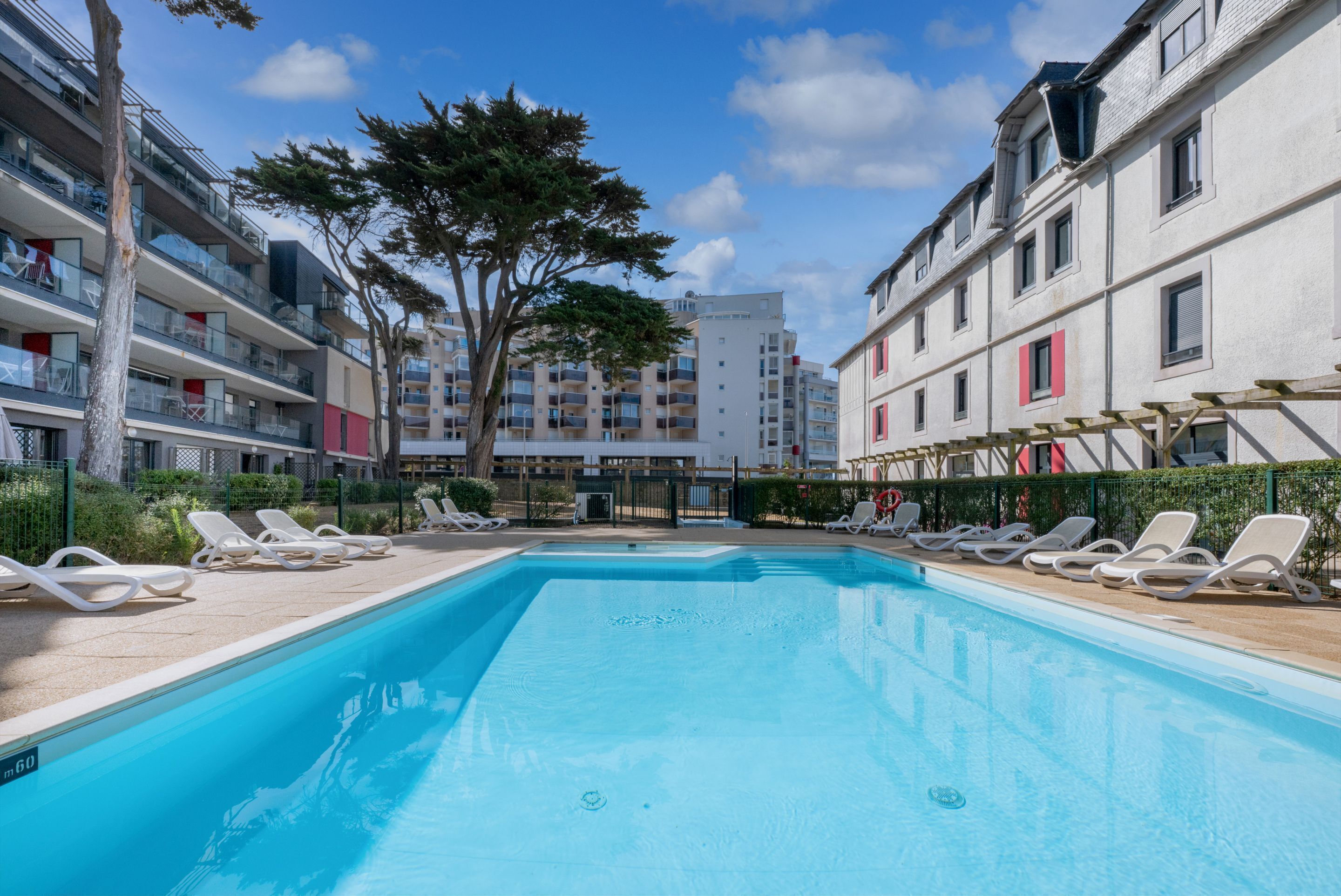 A swimming pool with lounge chairs, surrounded by multi-story buildings with balconies and a tree in the center.