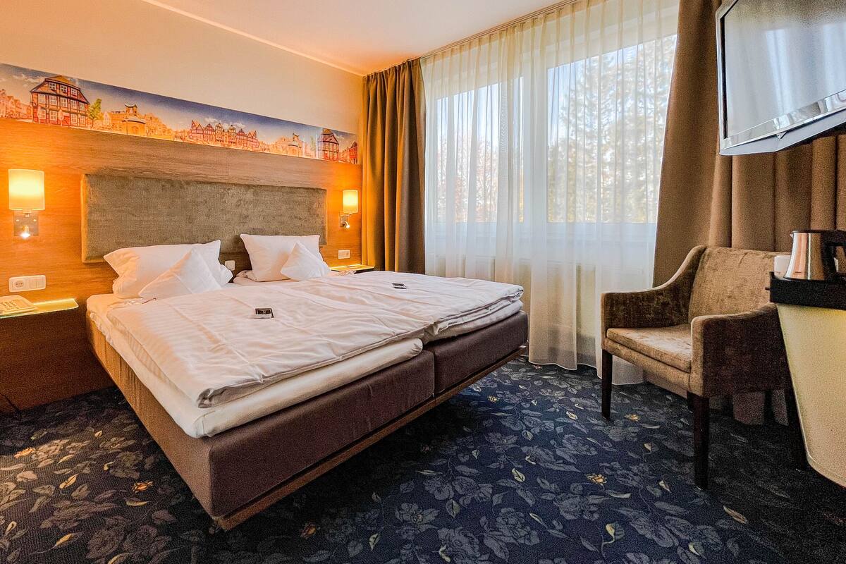 A hotel room with a large bed, a chair, a desk, and a painting of a cityscape on the wall.