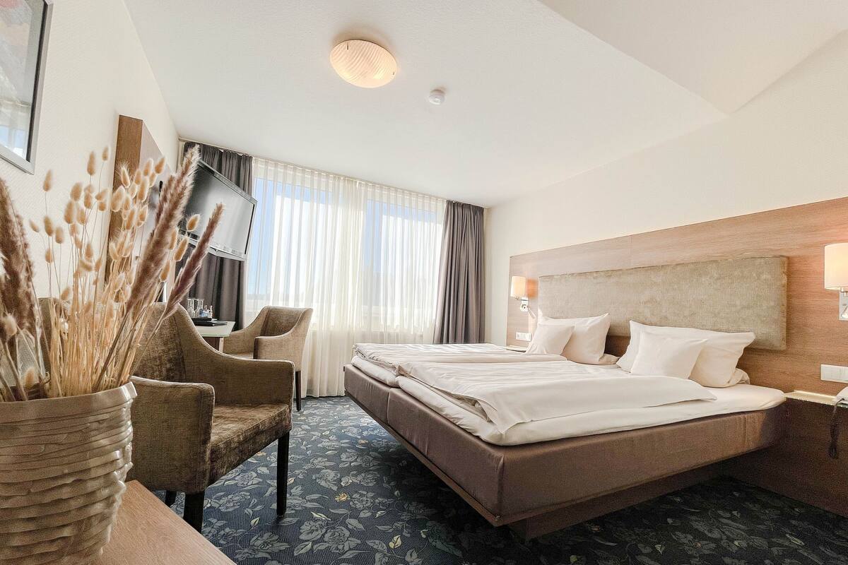 A hotel room with a large bed, a desk, a chair, and a vase with dried plants.