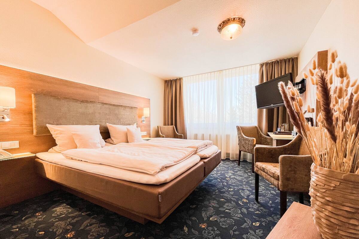 A hotel room with a large bed, a desk, a chair, a TV, and a decorative vase with pampas grass.