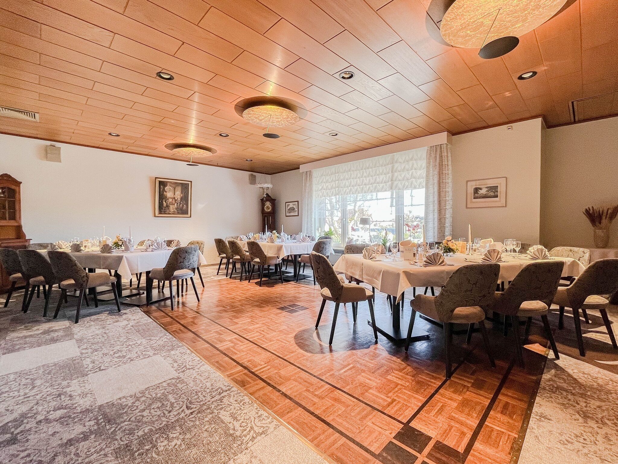 A formal dining room with multiple tables set for service, wooden flooring, and a large window providing natural light.