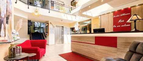 Vestíbulo de hotel con mostrador de recepción, asientos rojos, una escalera y vista al interior.