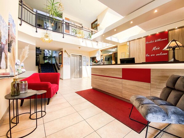 A hotel lobby with a reception desk, red seating, a staircase, and a view of the interior.