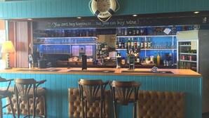 Una zona de bar con una pared azul, un letrero de bienvenida y un cartel que dice “No se puede comprar la felicidad…¡pero sí vino!”. Hay tres taburetes de bar y una barra con una caja registradora.