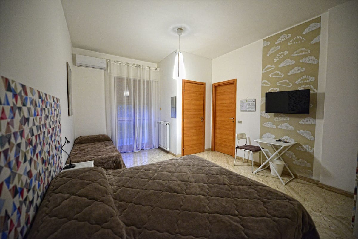 A room with two beds, a small table, a chair, a TV, and a patterned wall.