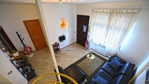 Una sala de estar con un sofá de cuero negro, una mesa de centro, una mesita auxiliar, una planta, una estantería, una puerta y una ventana con cortinas.