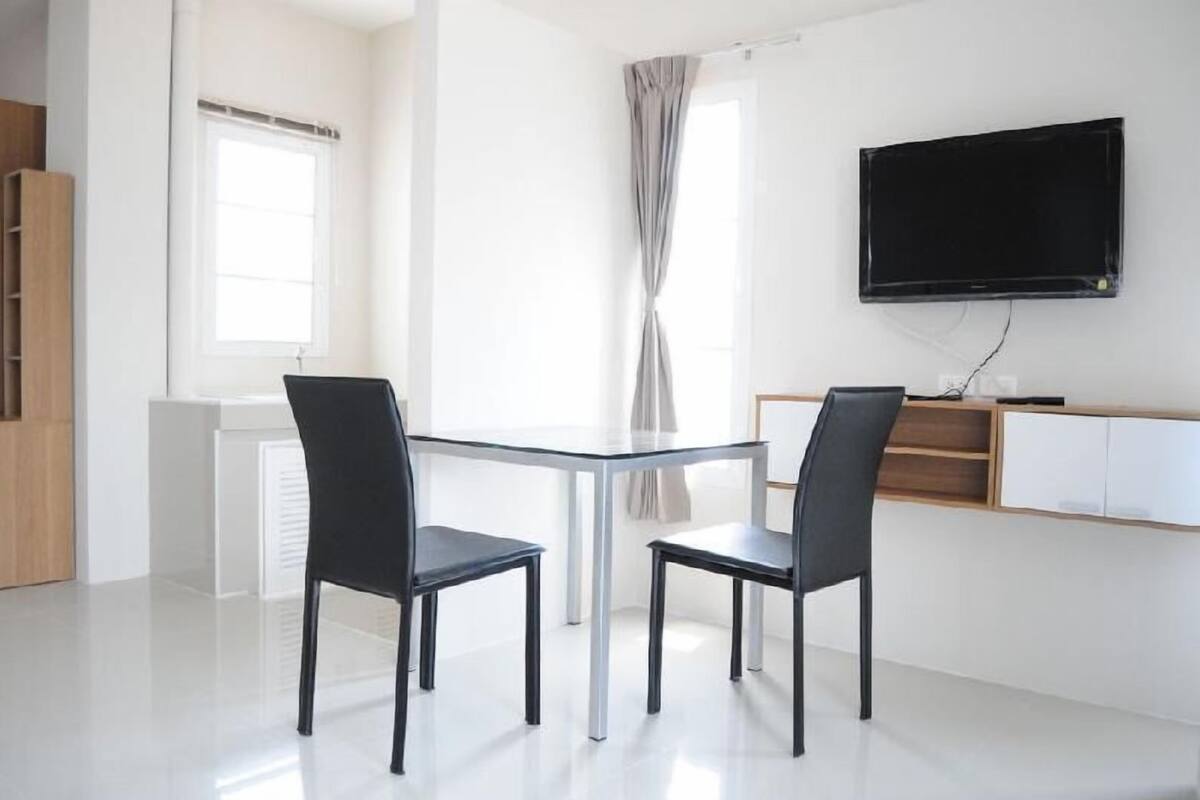 A modern room with a glass table, two black chairs, a TV mounted on the wall, and a window with curtains.
